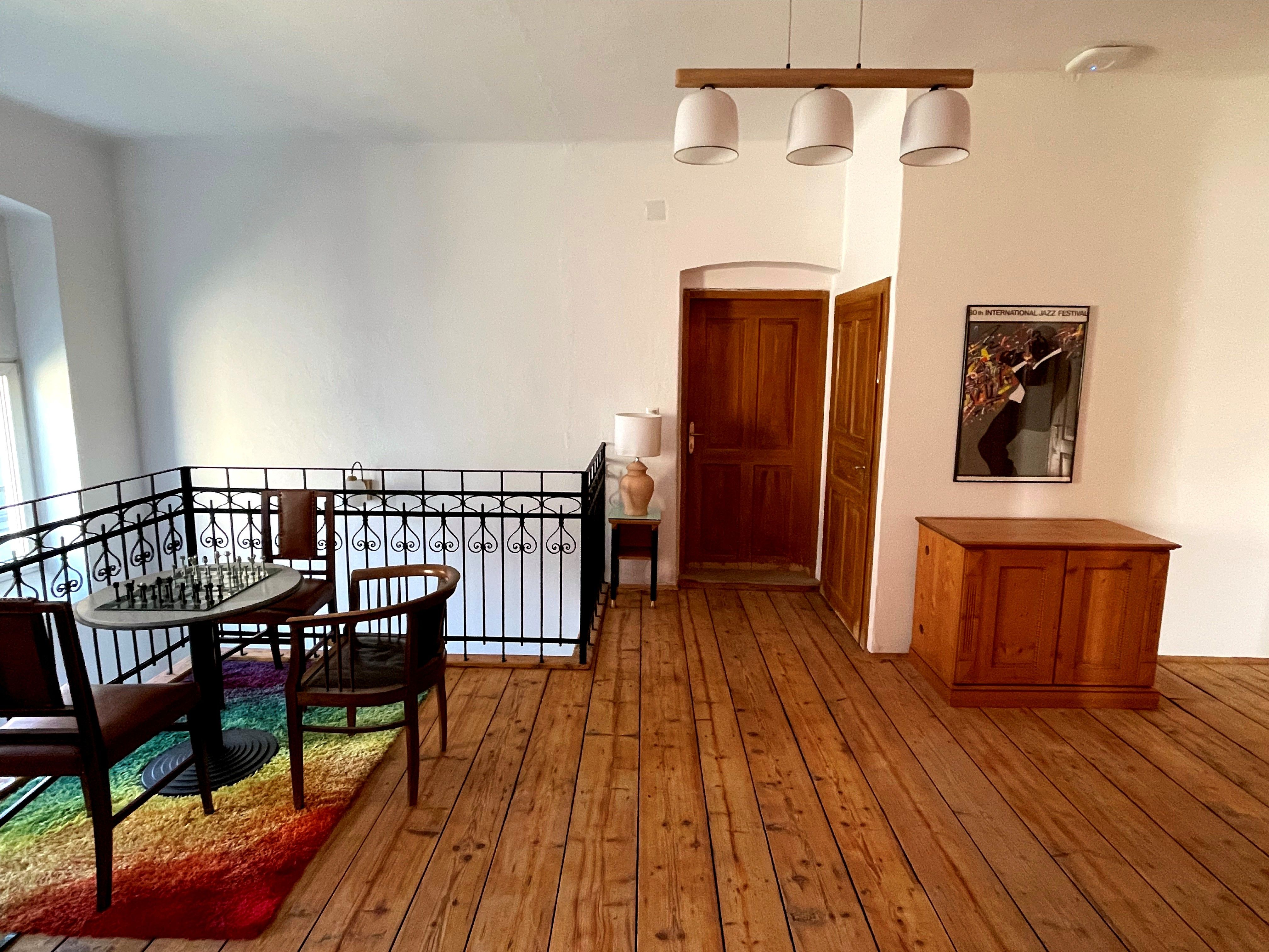 A cozy foyer with a wooden floor, a table with a chessboard, two chairs, and a colorful rug. A jazz festival poster hangs on the wall.