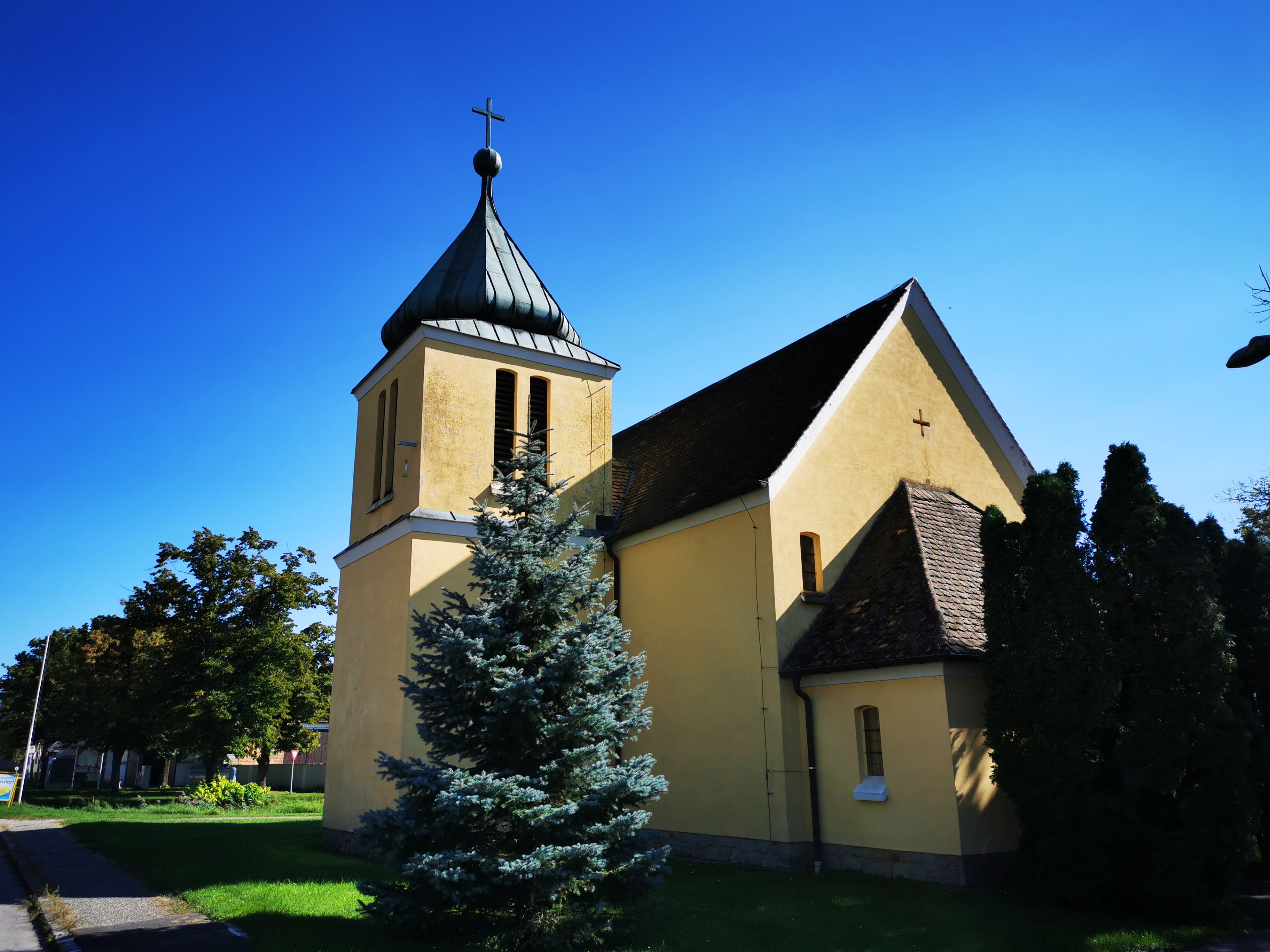 Yellow chapel with tower and cross, surrounded by trees, under a blue sky.