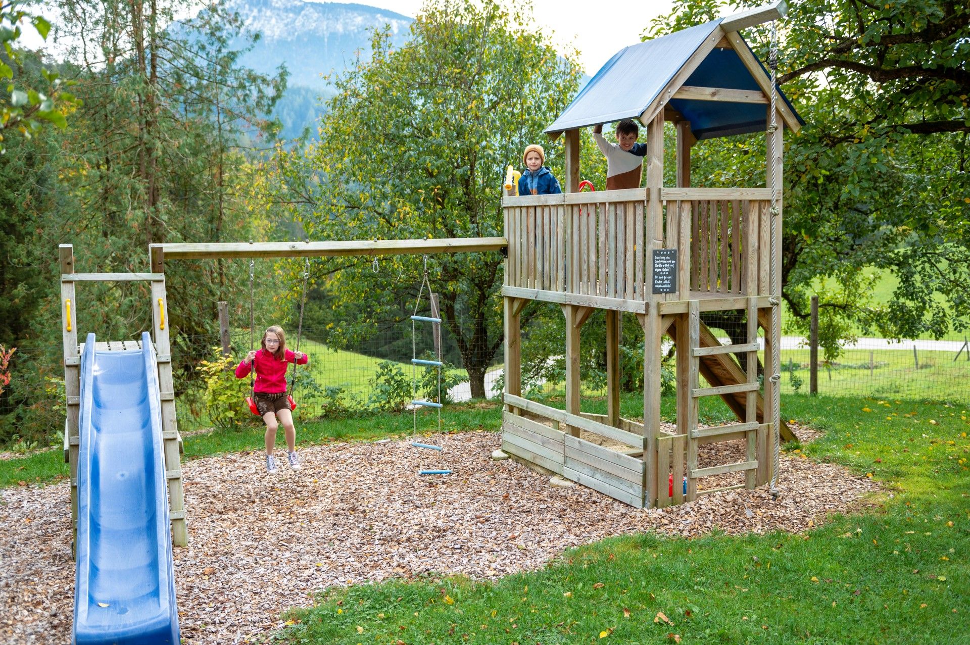 Children play on a playground with a swing, slide and climbing tower in a green setting.