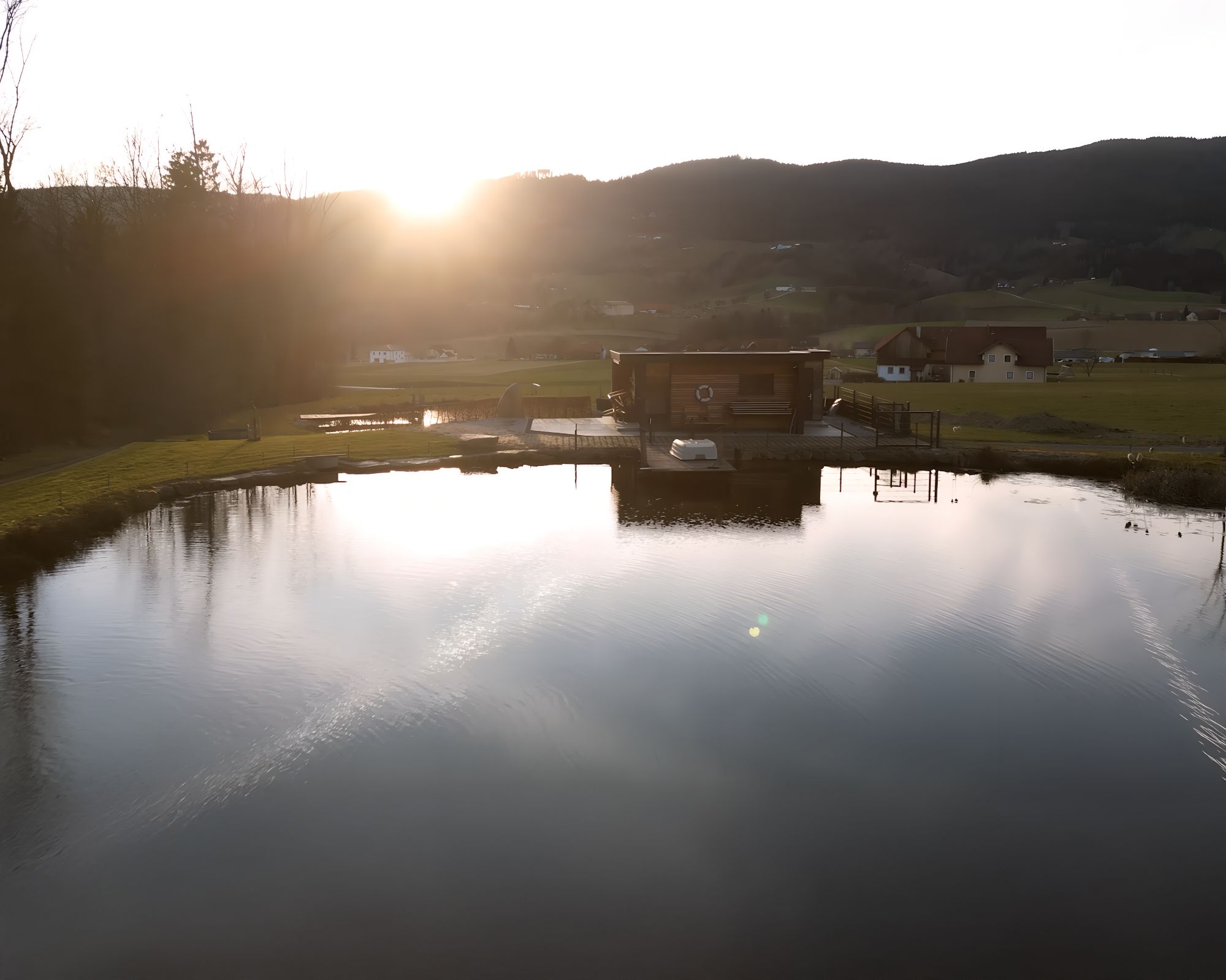 A pond with a wooden building on the shore, surrounded by hills and trees at sunset.