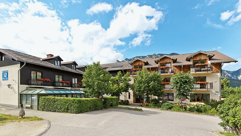 A traditional inn with wooden balconies and flowers, surrounded by trees and mountains under a blue sky.