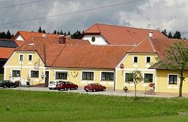Yellow building with red roofs and several cars in front of it, surrounded by green meadows and trees.