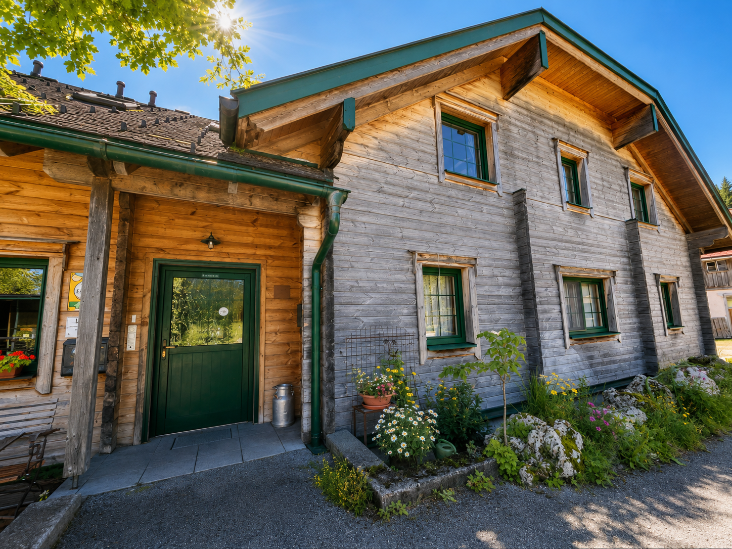 A traditional wooden house with green window frames and a green door, surrounded by flowers and plants, under a clear blue sky.