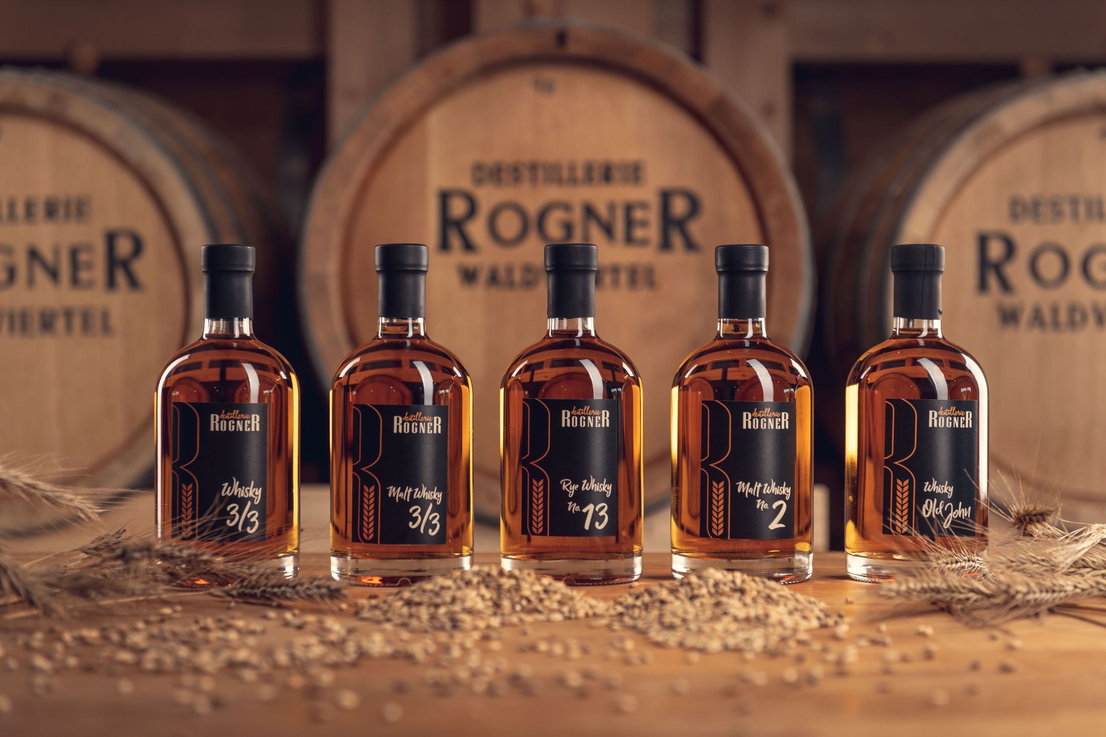 Five bottles of whisky from the Rogner distillery in front of wooden barrels.