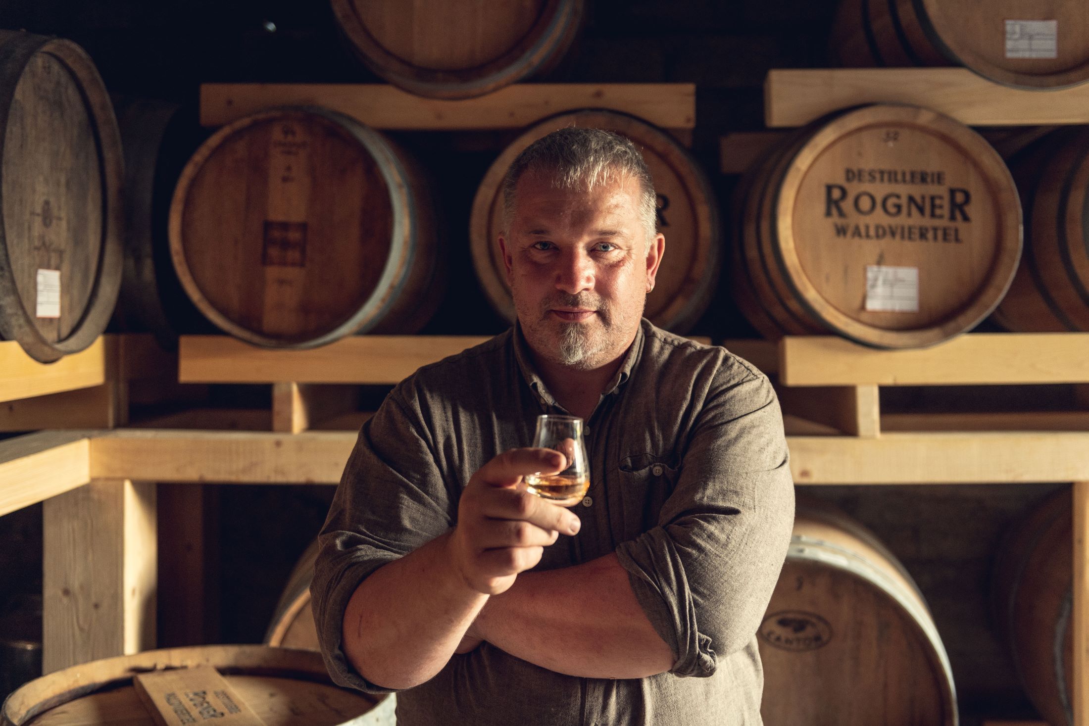 Man in a distillery with a glass in his hand in front of wooden barrels.