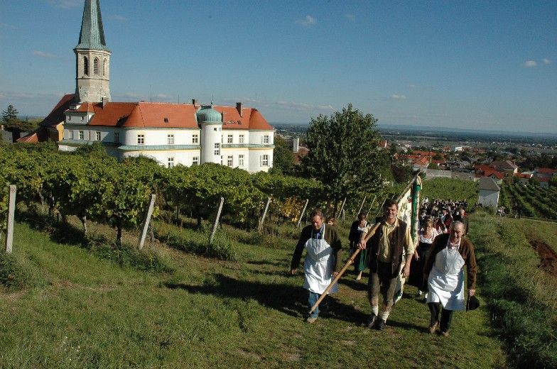 People in traditional dress walk through vineyards in Gumpoldskirchen, with a church and buildings in the background.