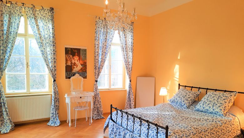 A cozy bedroom with a bed, blue floral patterns, a dressing table and a chandelier.