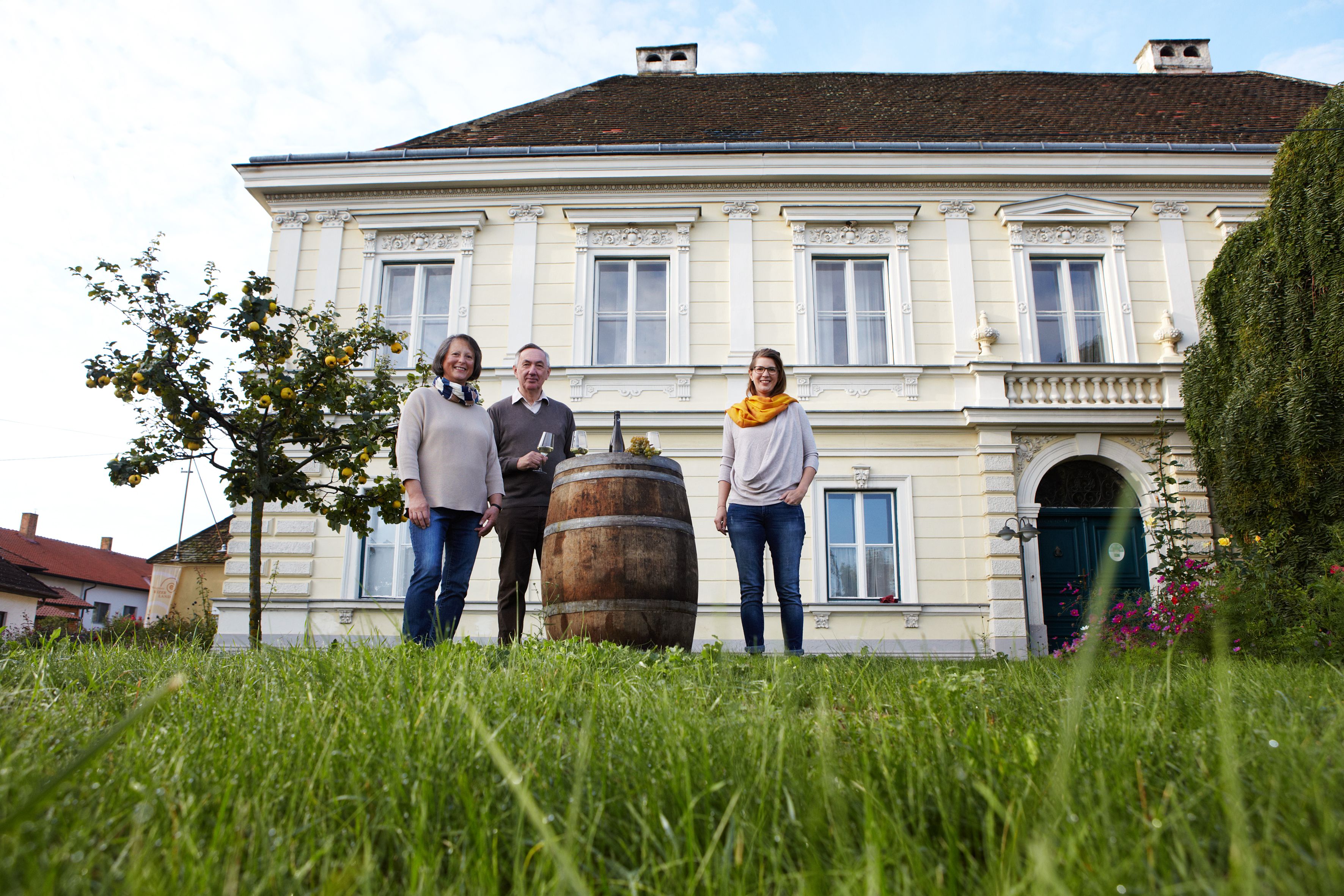 Three people stand in front of a historic building with a wine barrel in the foreground.