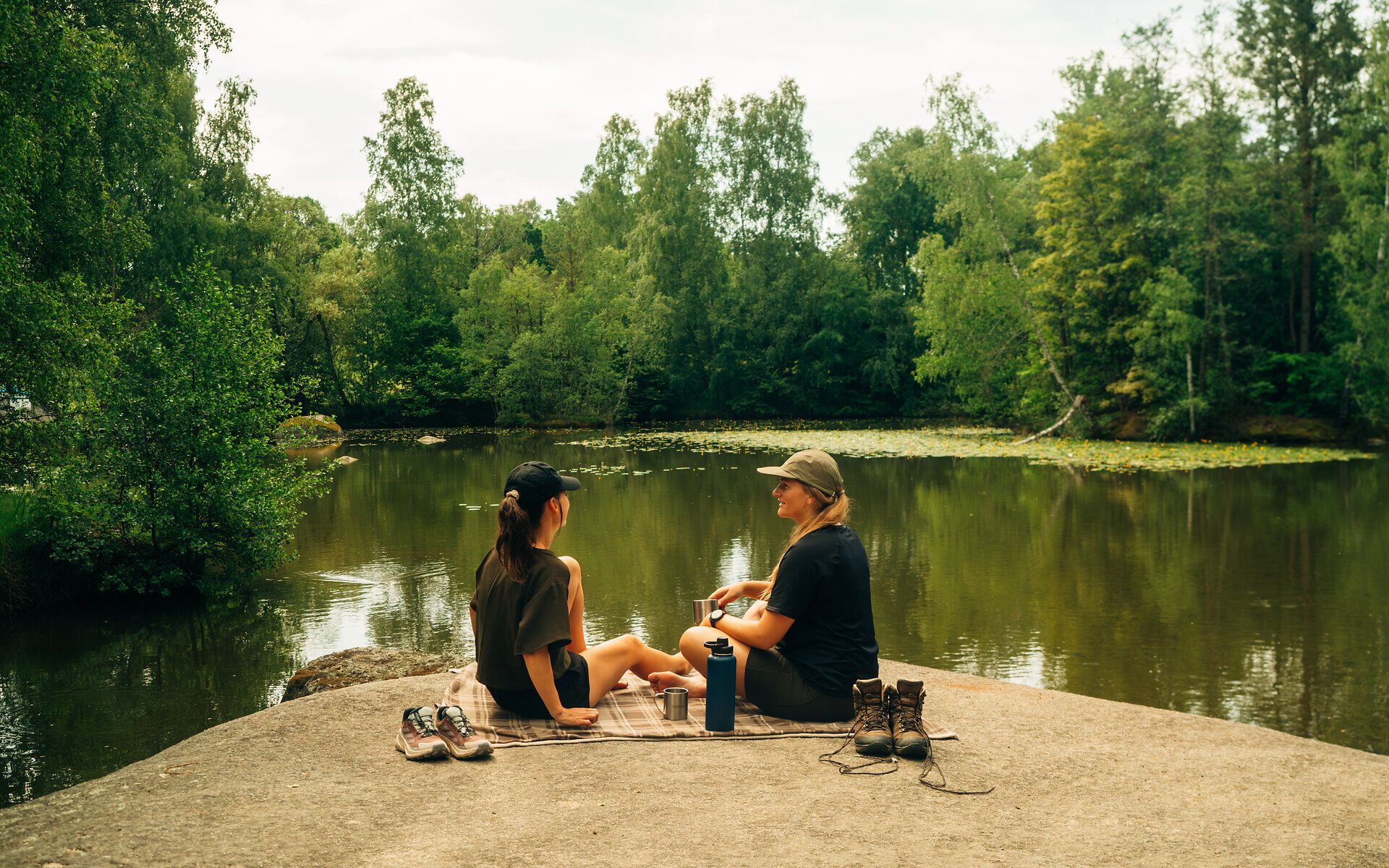 Two women sit on a stone in the block heath, surrounded by forest and water, taking a break from hiking