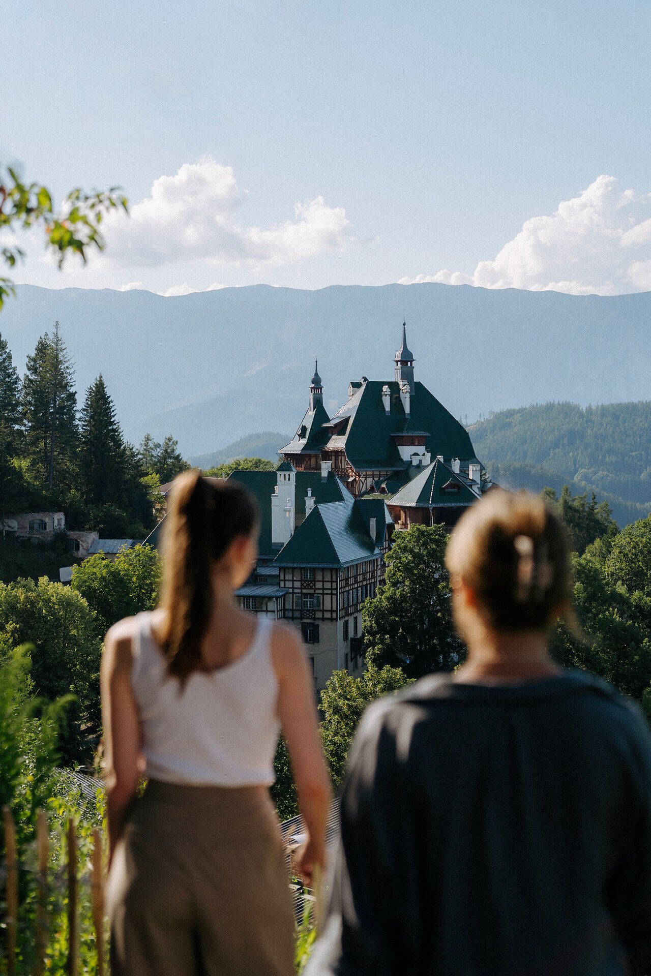 Two people, with their backs to the camera, look out over a picturesque mountain landscape. In the foreground you can see the Südbahnhotel, nestled in lush greenery. In the background, mountains rise up under a clear blue sky.