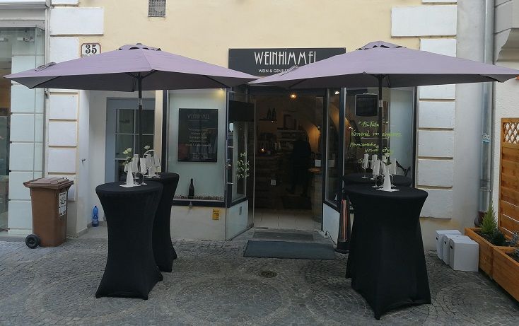 Entrance to a wine bar with two bar tables and parasols in front of it.