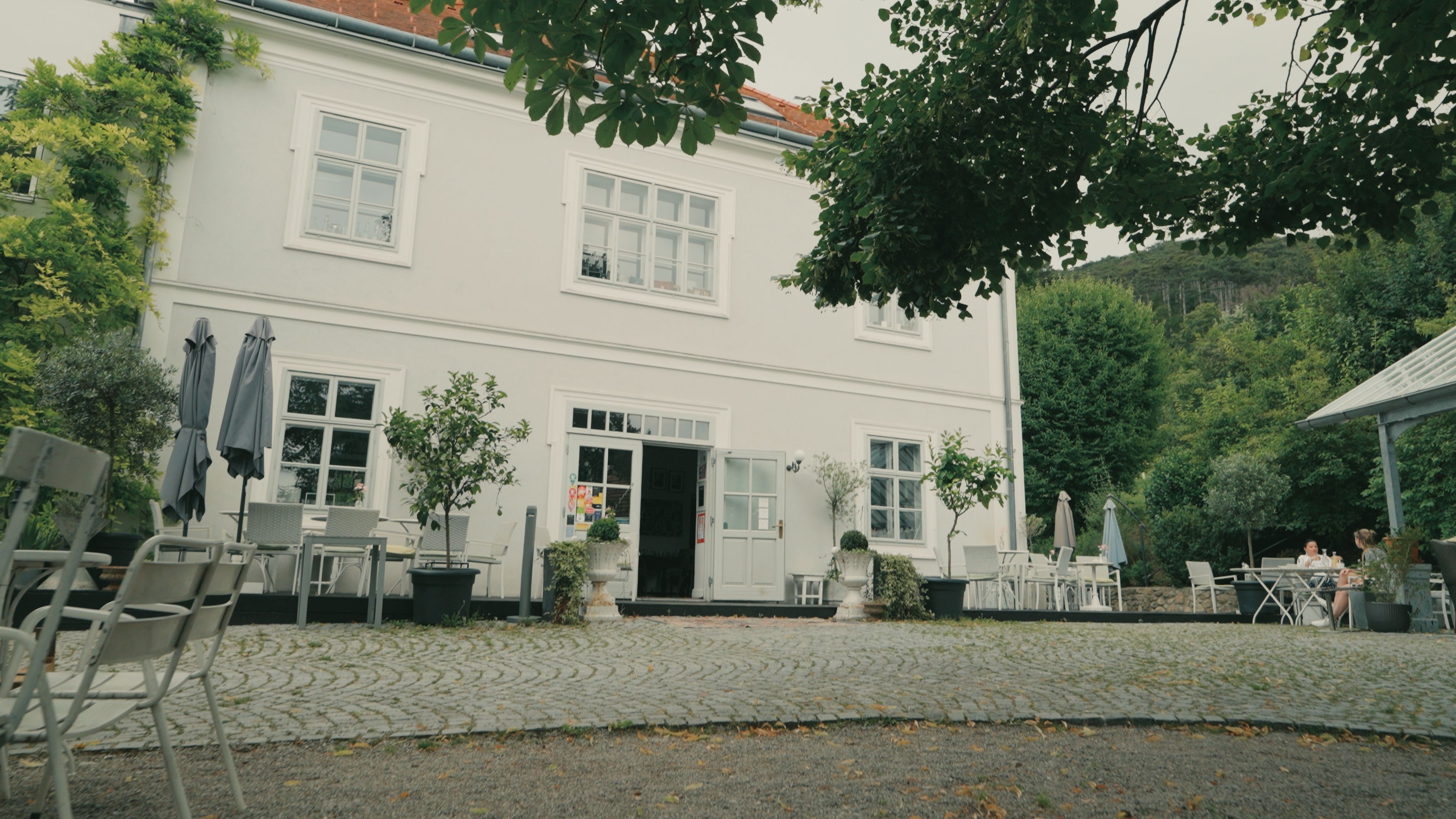 A white historic building with an open entrance door, surrounded by deciduous trees