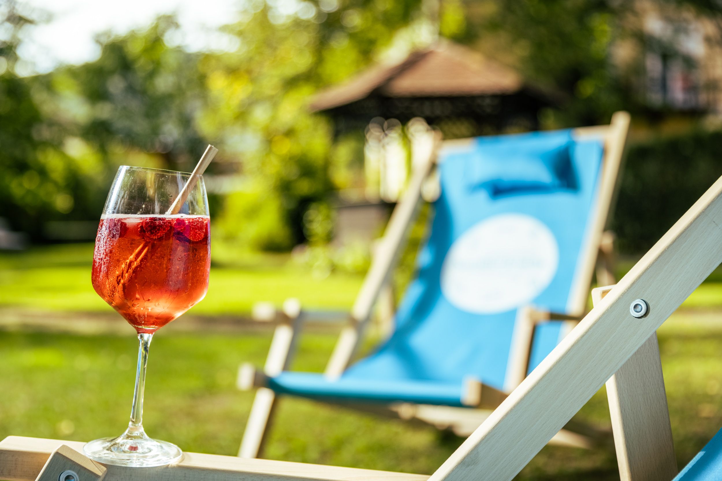 A glass with a red drink and a straw on a deckchair in the garden.