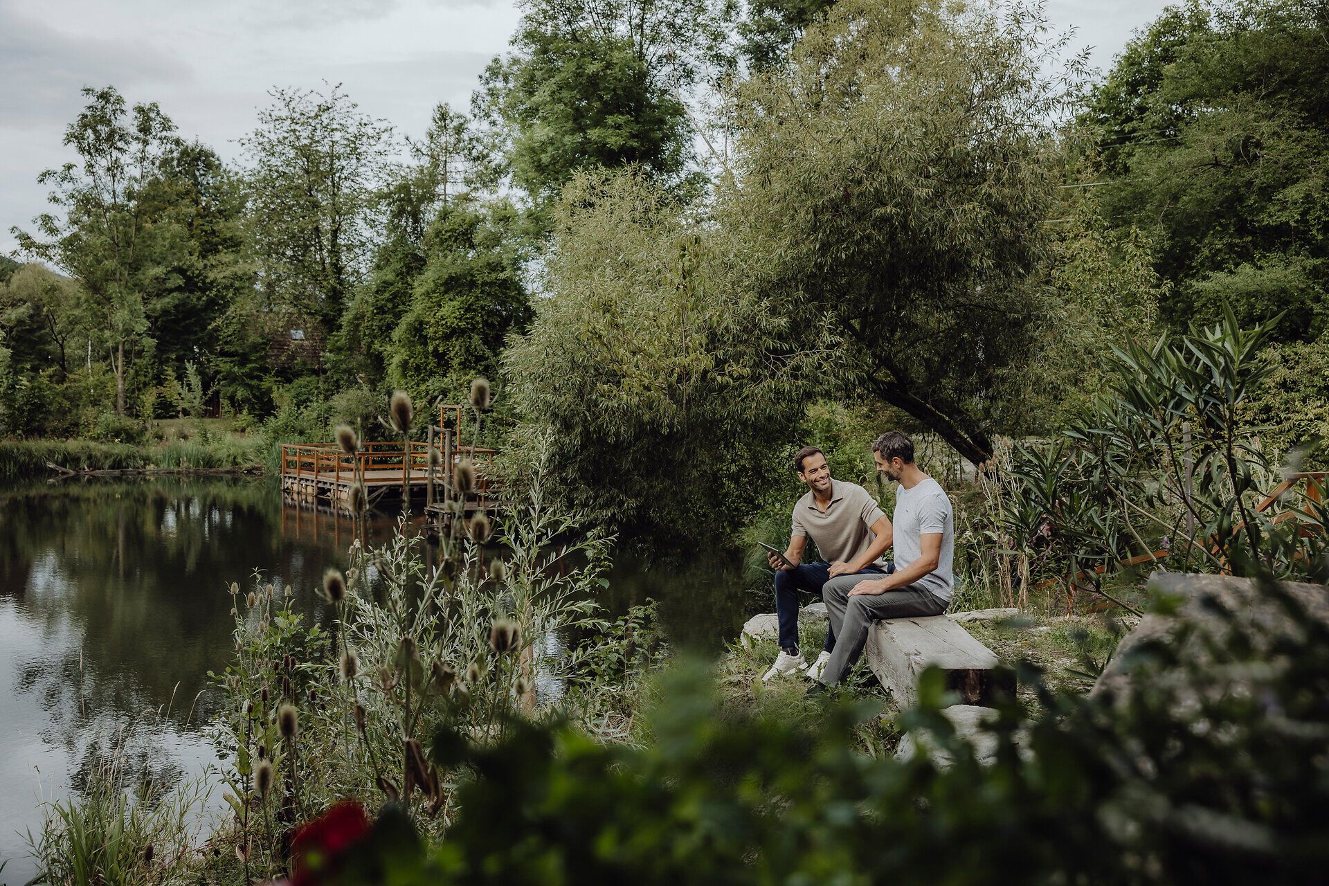 Inmitten einer idyllischen Landschaft am Ufer eines ruhigen Gewässers sitzen zwei Freunde entspannt auf einer Holzbank. Umgeben von üppigem Grün und blühenden Pflanzen genießen sie die friedliche Atmosphäre und die Schönheit der Natur.