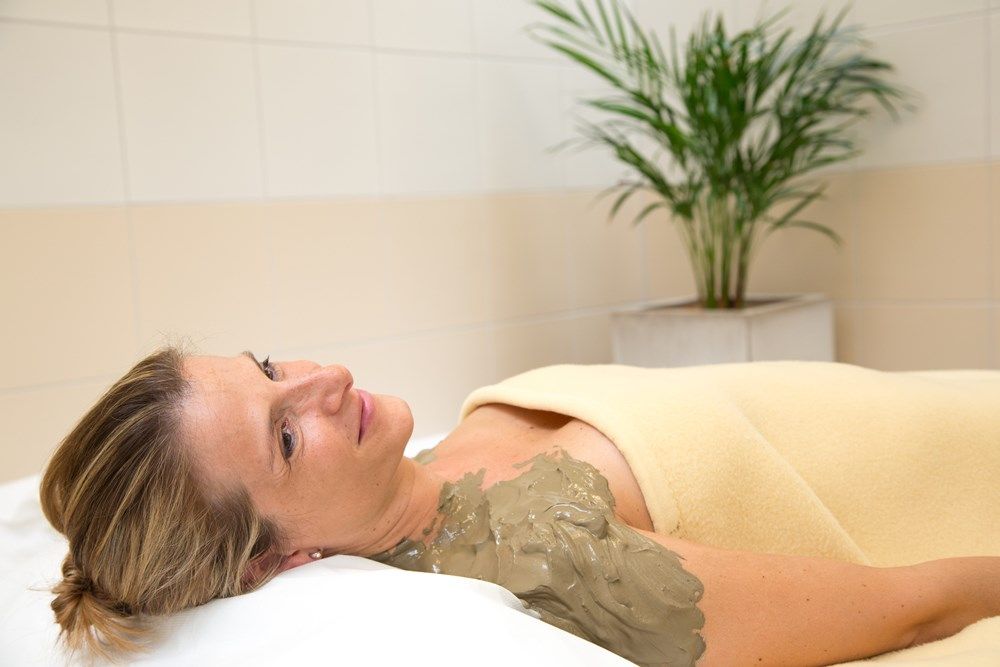 Woman relaxing with a sulphur mud pack on her shoulder, wrapped in a blanket, in the wellness area with a plant in the background.