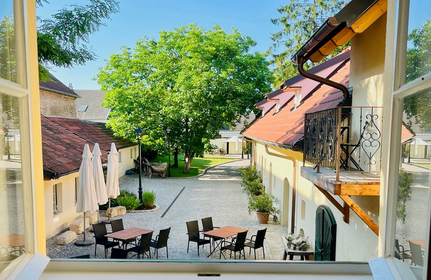 View from a window onto an inner courtyard with tables, chairs and parasols, surrounded by buildings and trees.