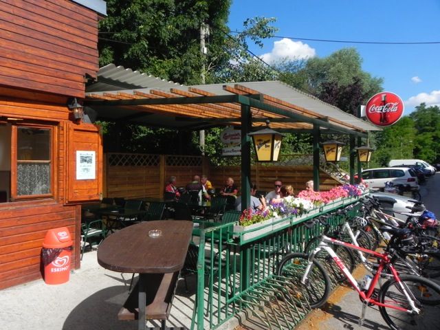 Outdoor area of a snack bar with wooden walls, tables and bicycles. People sitting under a covered area. Coca-Cola sign visible.