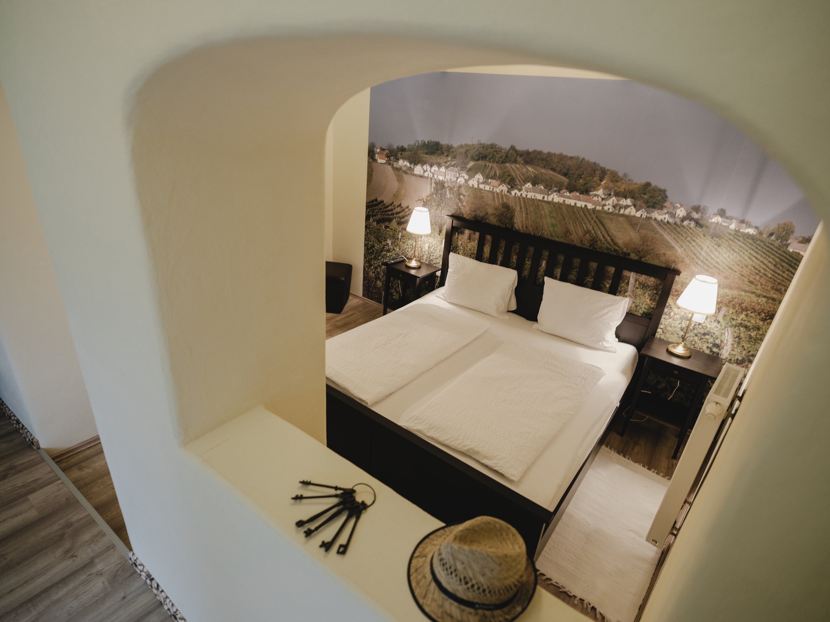 A cozy bedroom with a double bed, two bedside lamps and a wallpaper with a wine landscape.