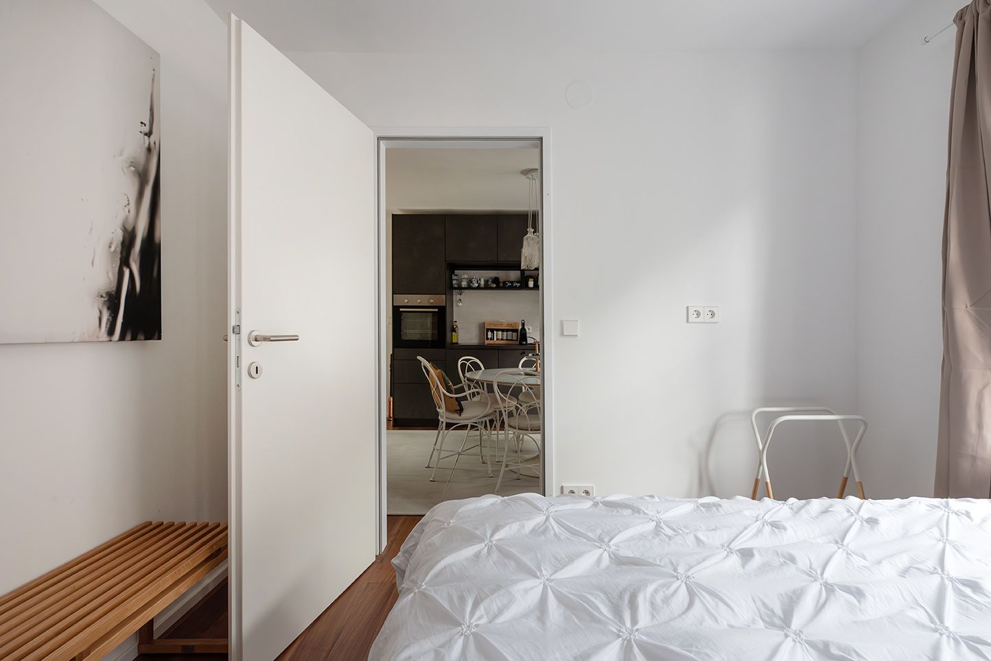 A modern bedroom with white bed linen, an open door leading to a dining area and minimalist furnishings.