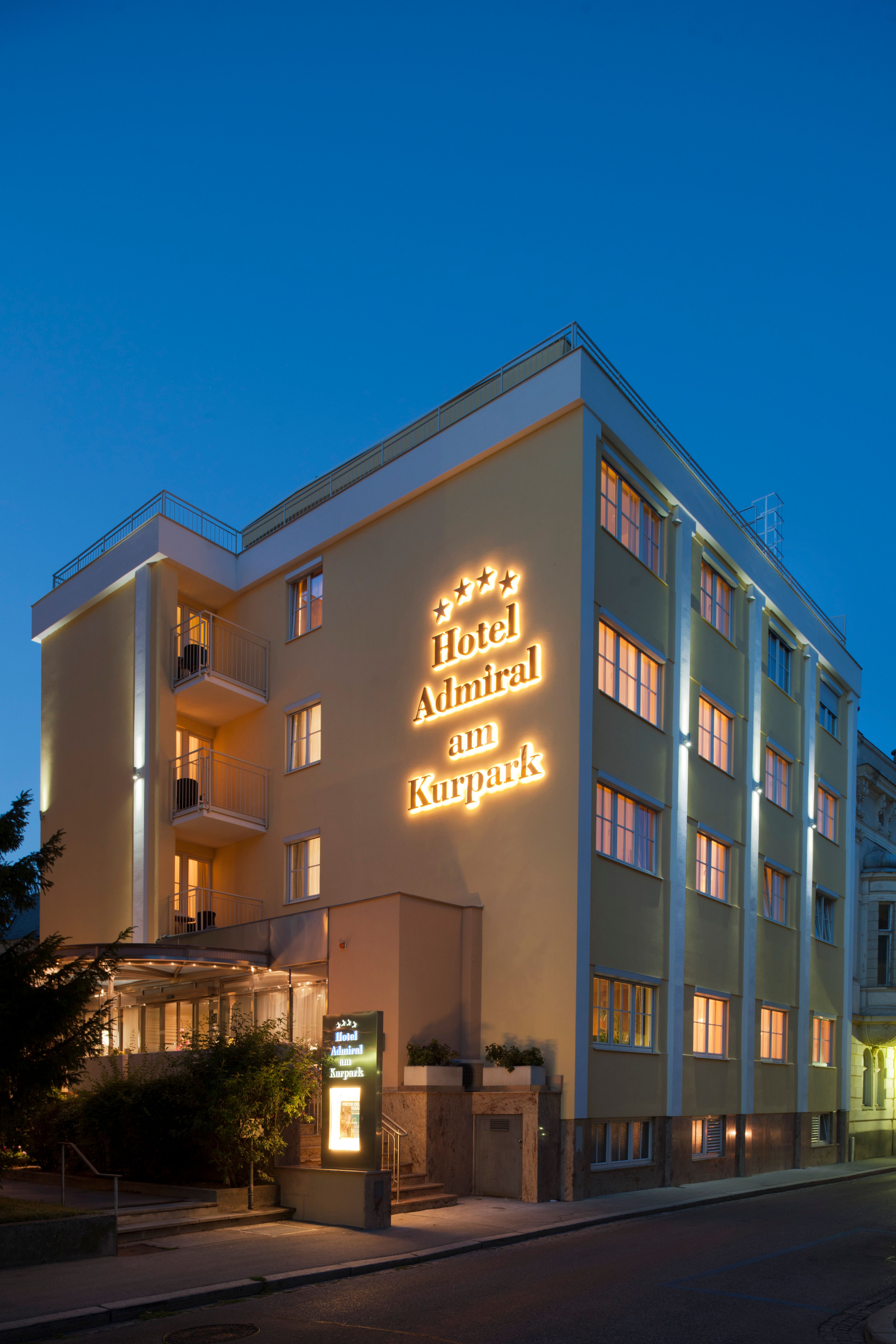 Exterior view of a hotel at dusk with illuminated lettering "Hotel Admiral am Kurpark".