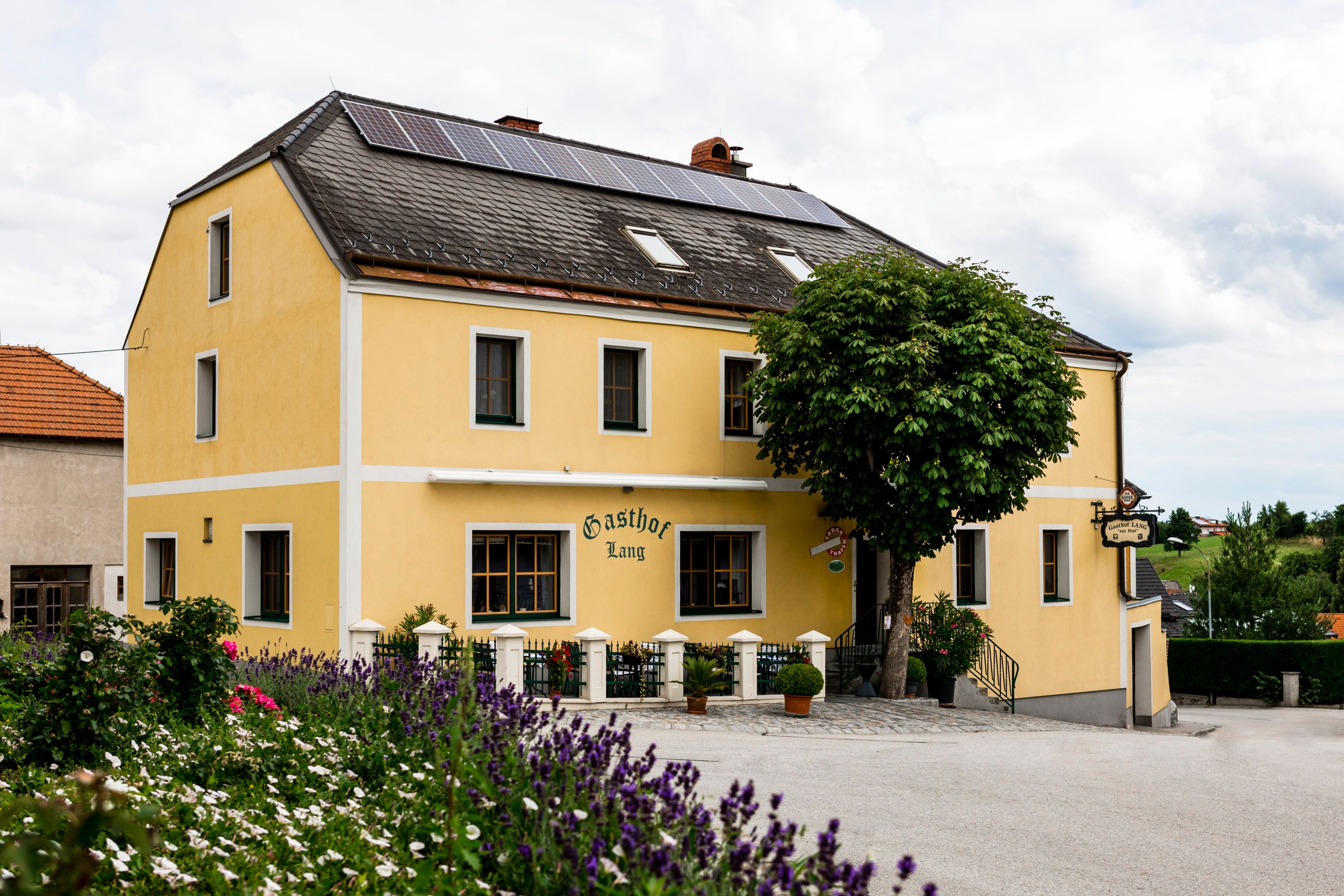 Yellow guesthouse with solar panels on the roof, surrounded by flowers and trees.