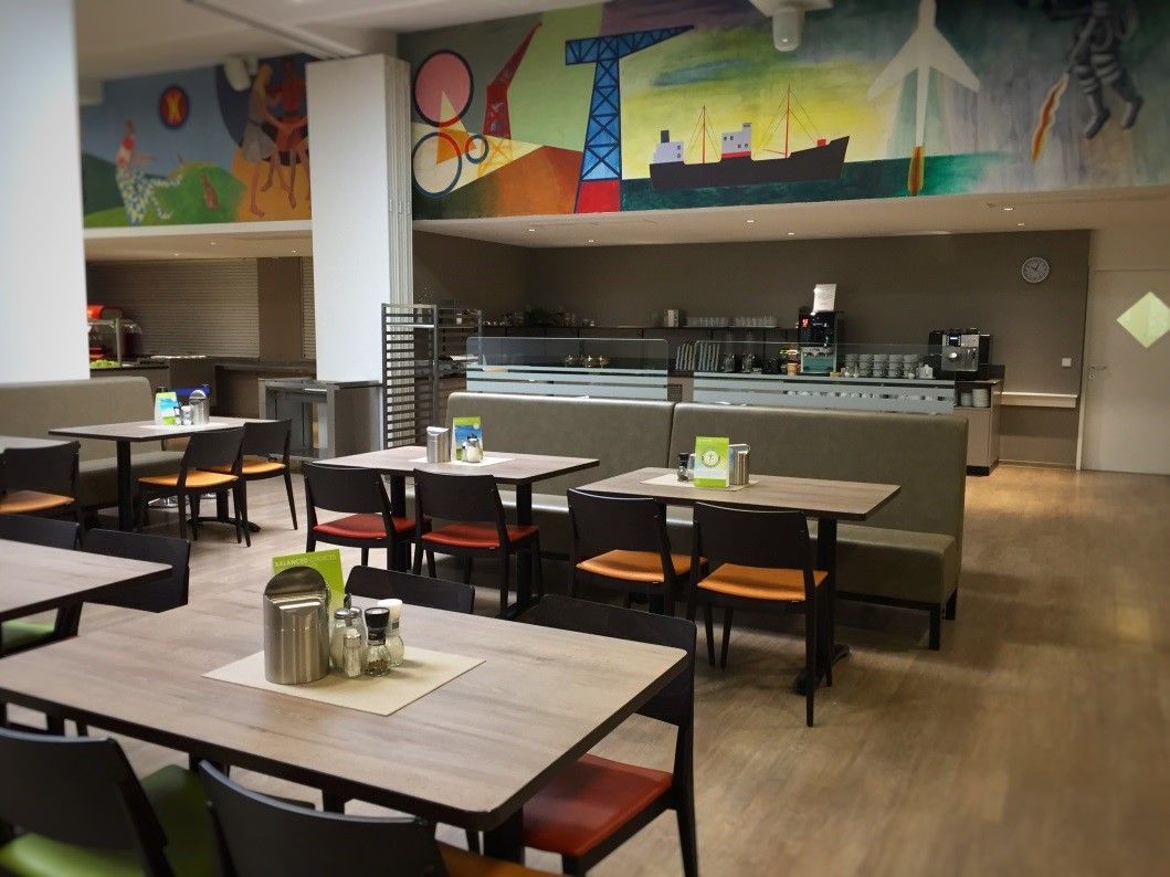 Interior view of a restaurant with empty tables and chairs, colorful wall painting in the background.