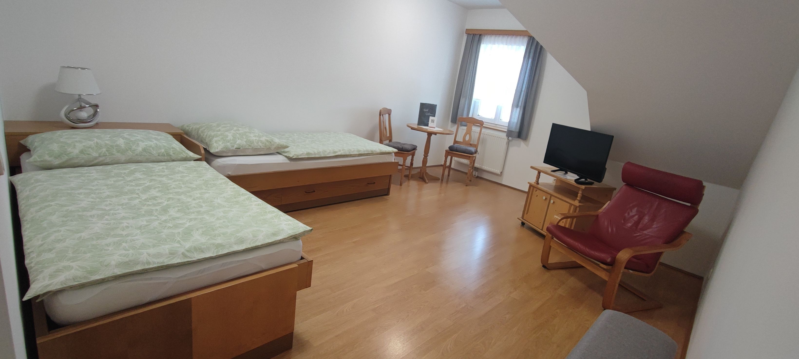 A simply furnished room with two single beds, a small table and chairs, a TV and a red armchair.