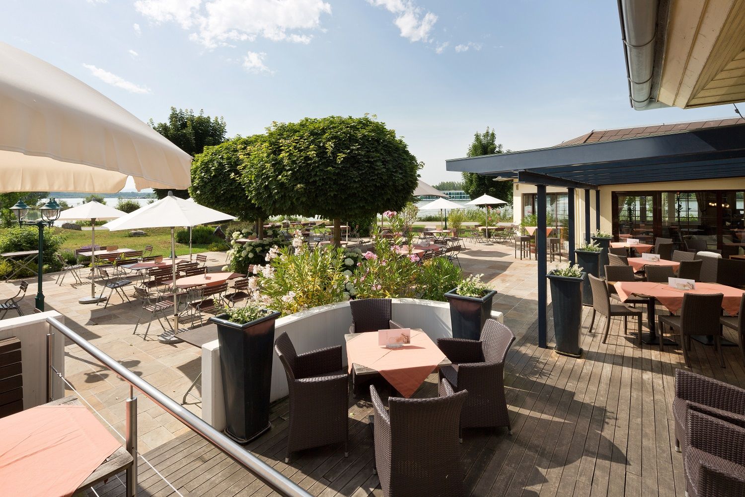 Sunny terrace with tables, chairs and parasols, surrounded by plants and trees.