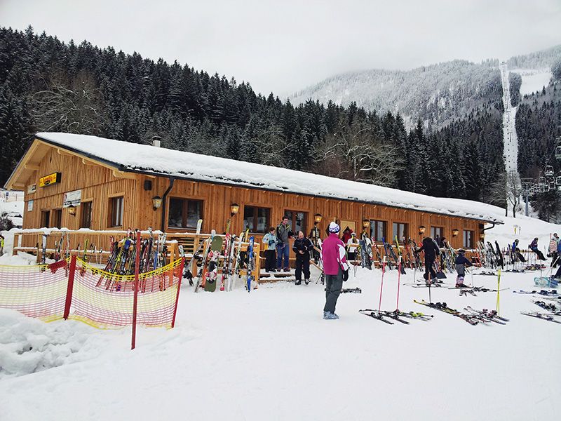 Ski hut in the snow with skiers and equipment in front of it.