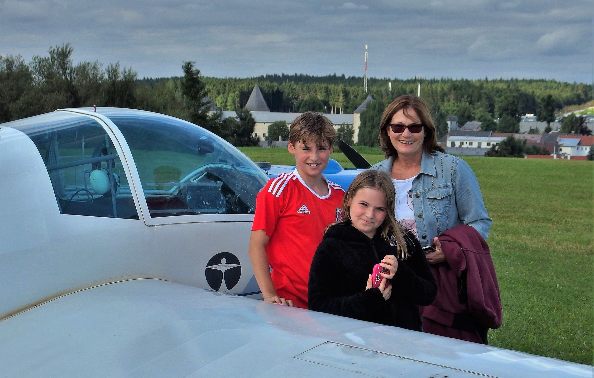 Three people are standing next to a small airplane on an airfield with a green meadow and a cloudy sky in the background.