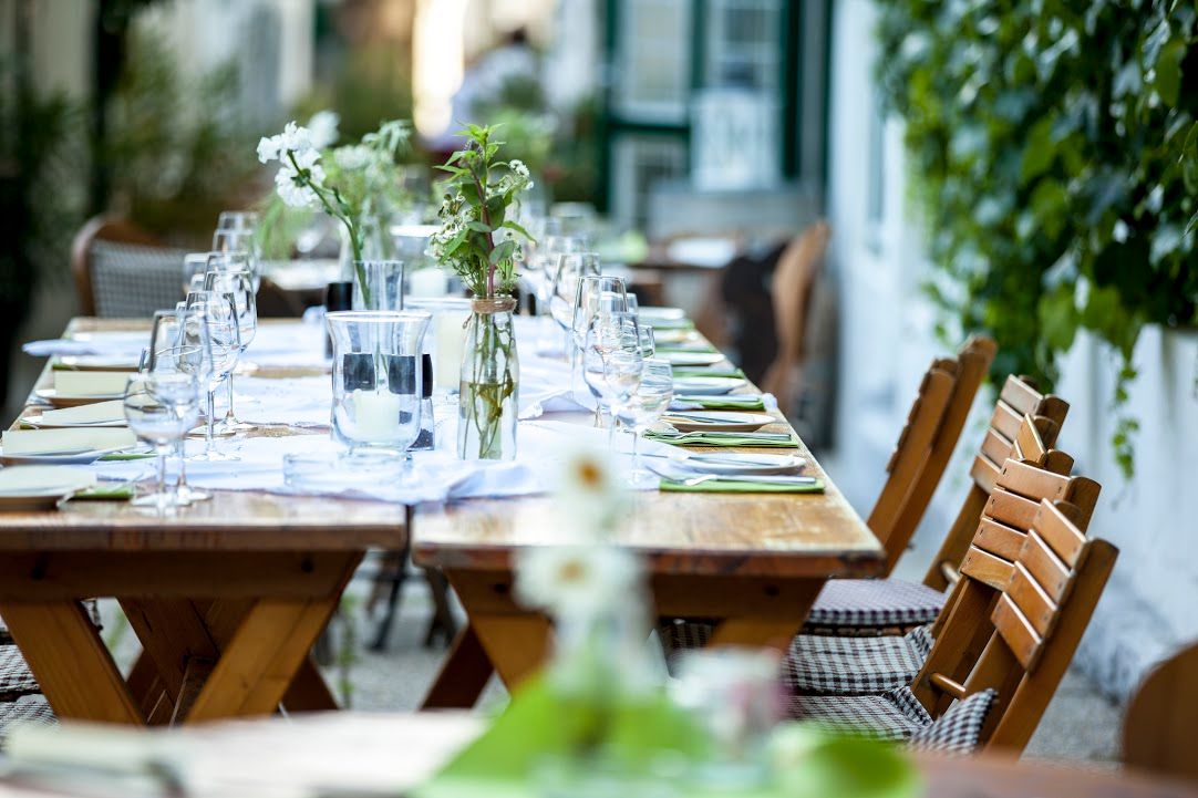 Long wooden table outside, set with glasses, cutlery and flowers.