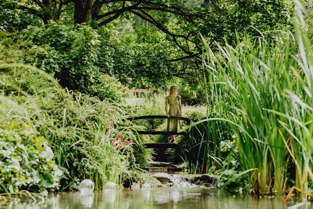 Woman standing on a wooden bridge in a lush, green garden with a stream.