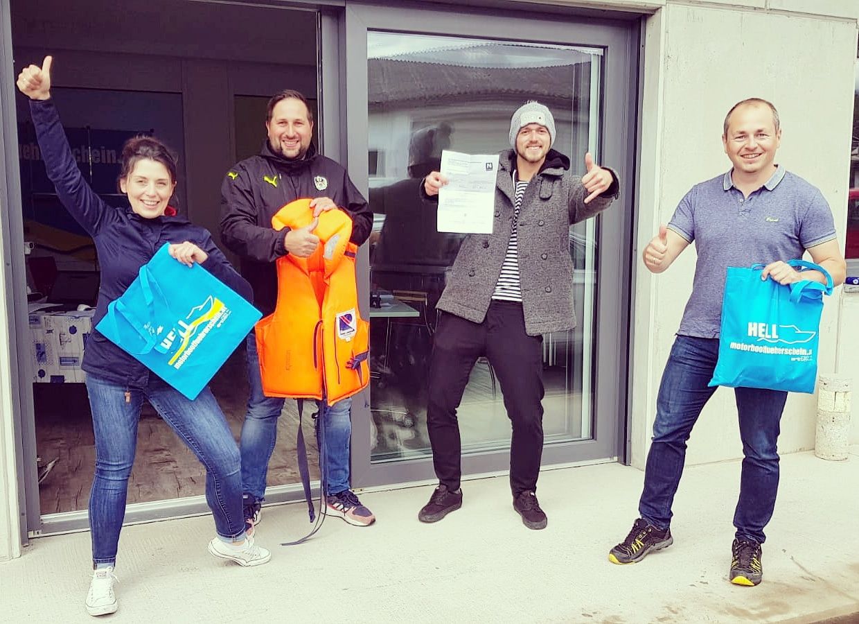 Four people are standing in front of a building, holding bags and a life jacket, smiling and giving the thumbs up.