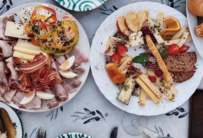 Two plates with cold cuts, cheese, vegetables and fruit on a table.