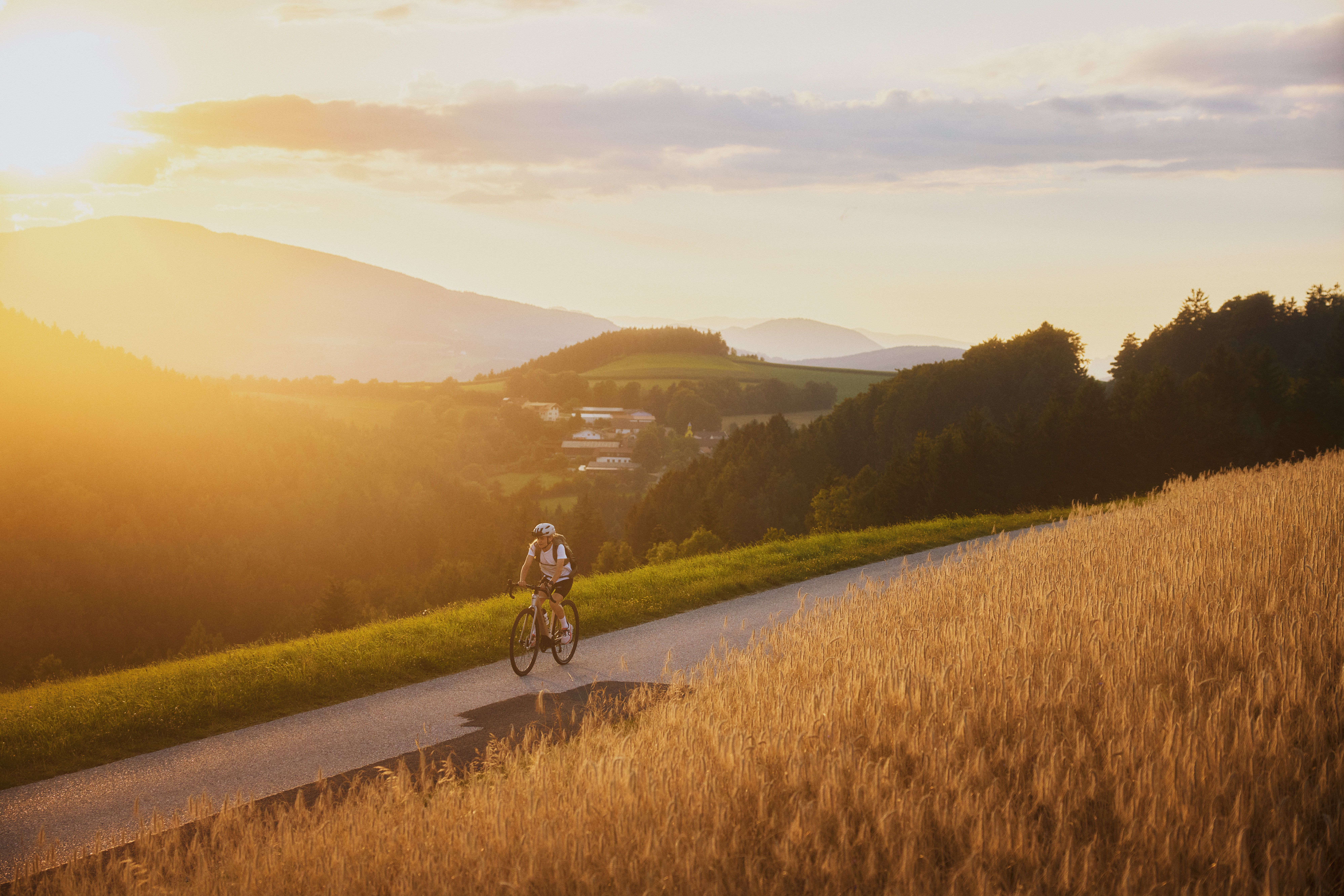 A cyclist rides along a country road at sunset through a hilly landscape of fields and forests.
