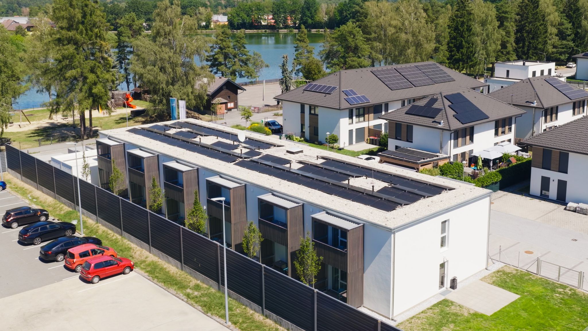 Aerial view of modern buildings with solar panels on their roofs, surrounded by trees, with a lake in the background.