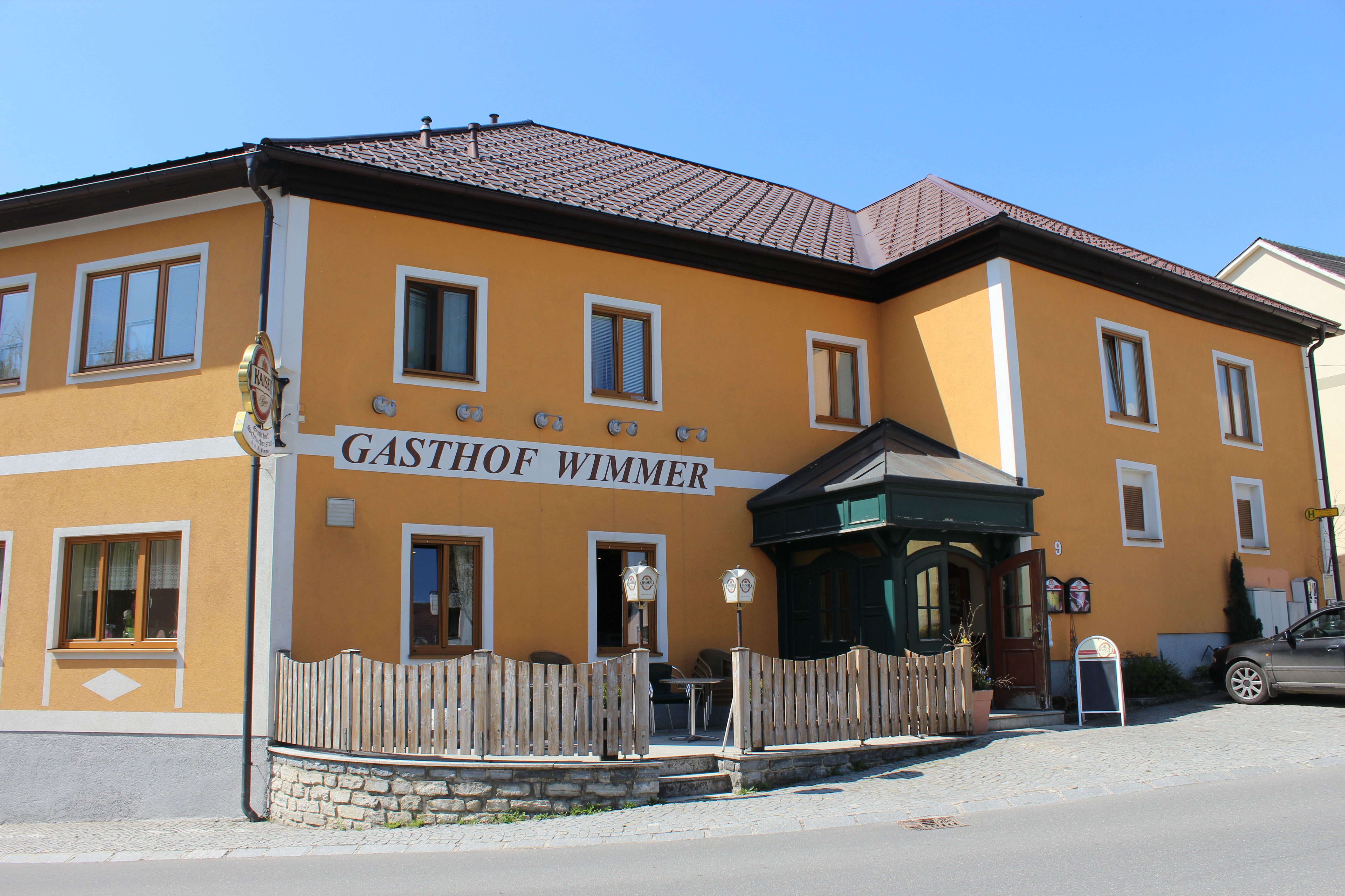 Yellow building with the inscription 'Gasthof Wimmer', wooden fence and entrance door.