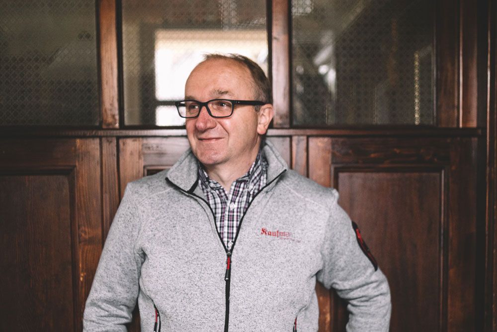 A man with glasses and a gray jacket stands in front of a wooden wall.