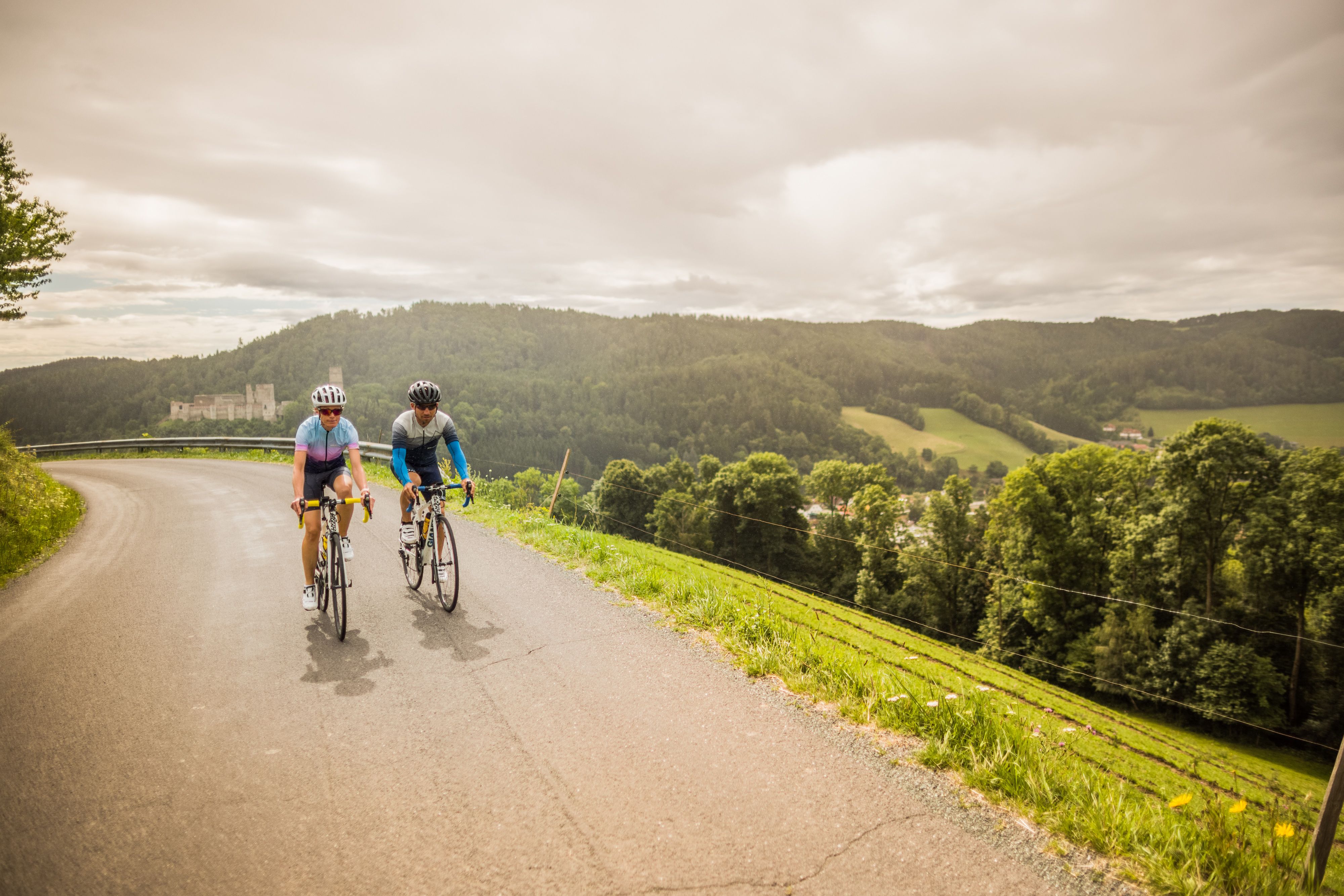 Two cyclists on a winding road in a hilly landscape with the ruins of Kirchschlag Castle in the background.