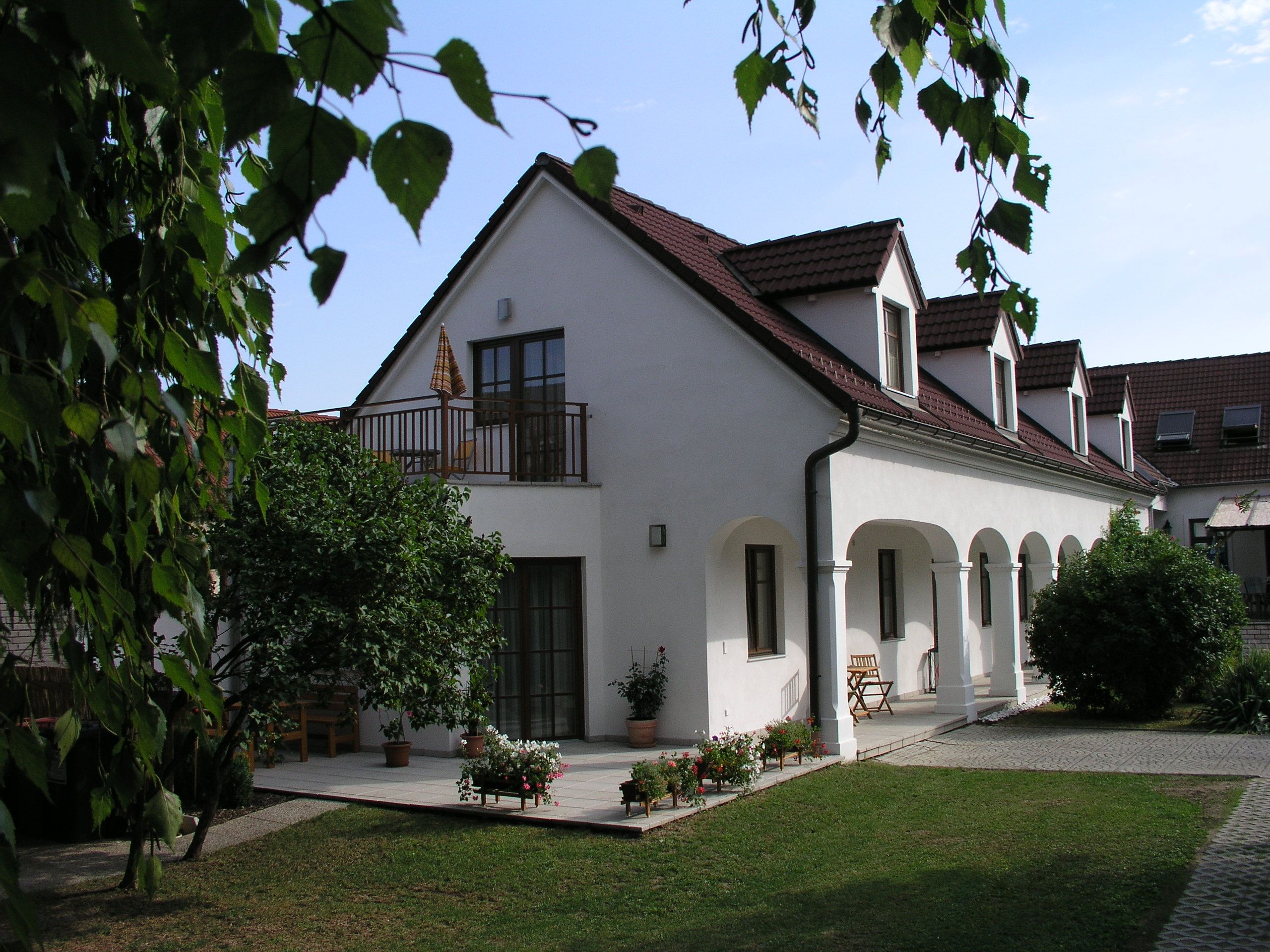 A white building with red roof tiles and arcades, surrounded by a garden with trees and flowers.