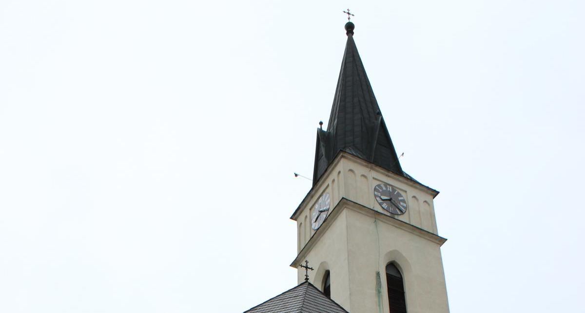 Church tower with clock and pointed roof in front of a bright sky.