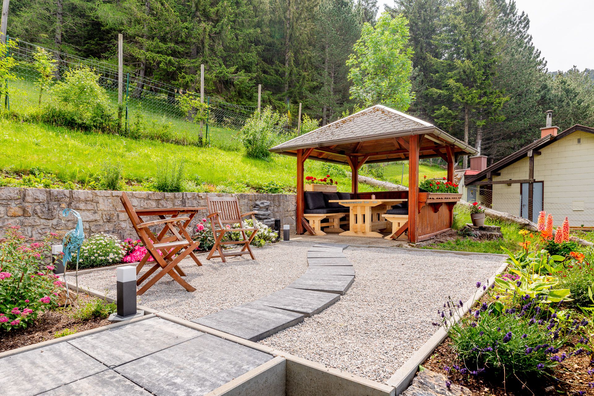 Garden with pavilion, wooden furniture and flower beds in front of a forest.
