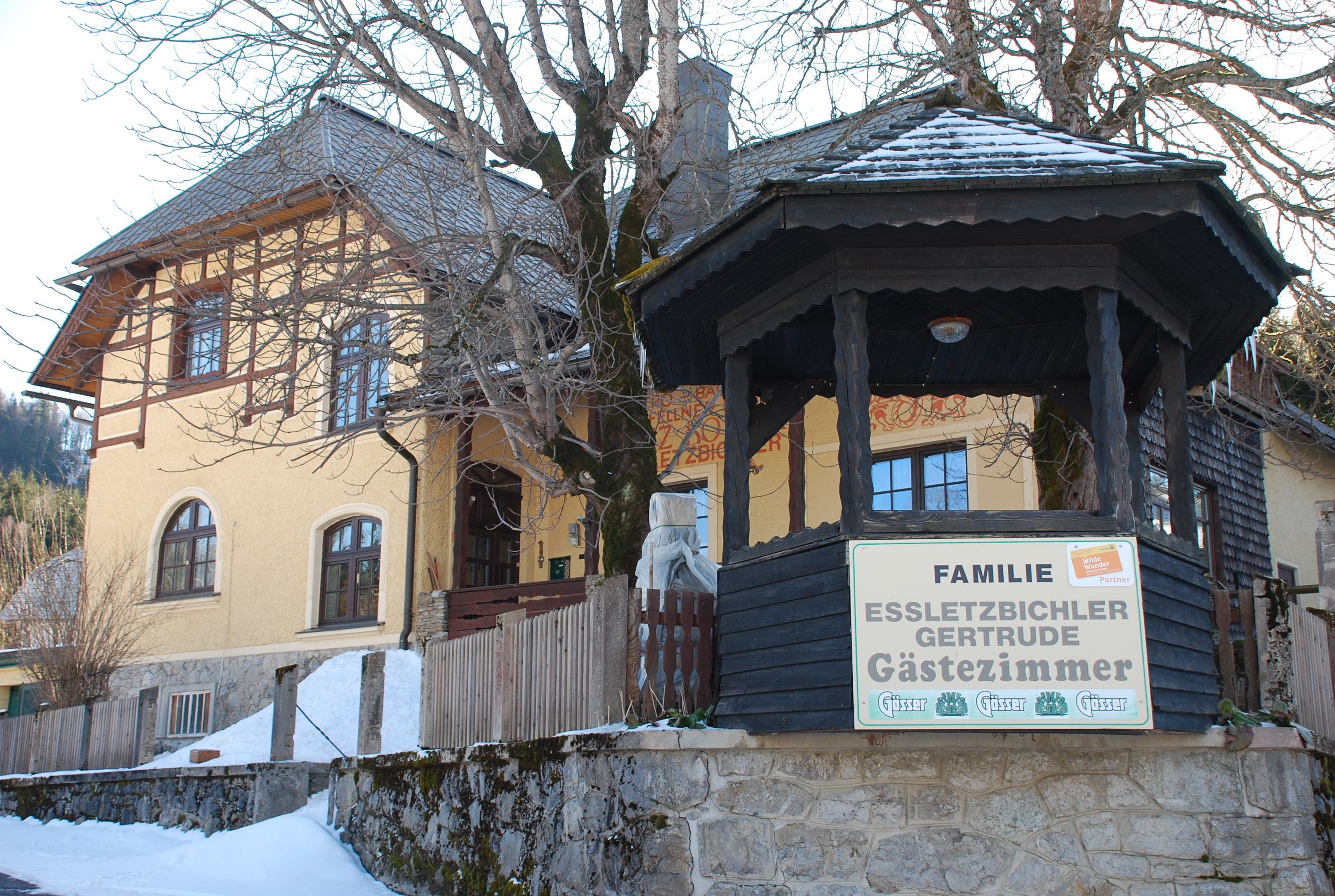 Yellow guest house with wooden veranda and sign 'Familie Essletzbichler Gertrude Gästezimmer'.