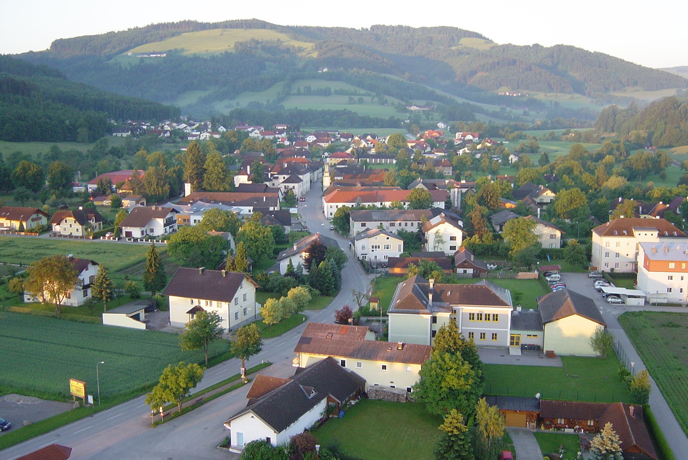 Aerial view of a village with houses, streets and surrounding hills in the background.