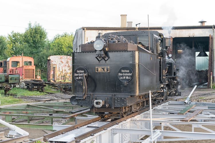 Steam locomotive in front of a boiler house with rails in the foreground.