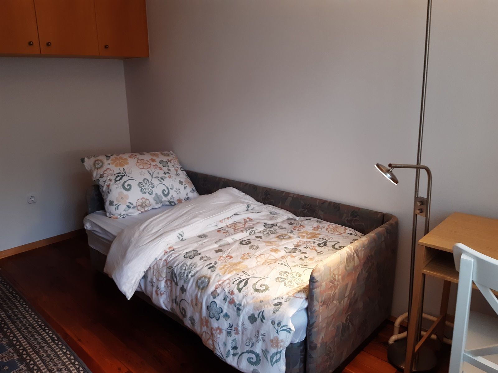 A small room with a single bed, a desk with a lamp and a chair. The bed has colorful floral bedding.