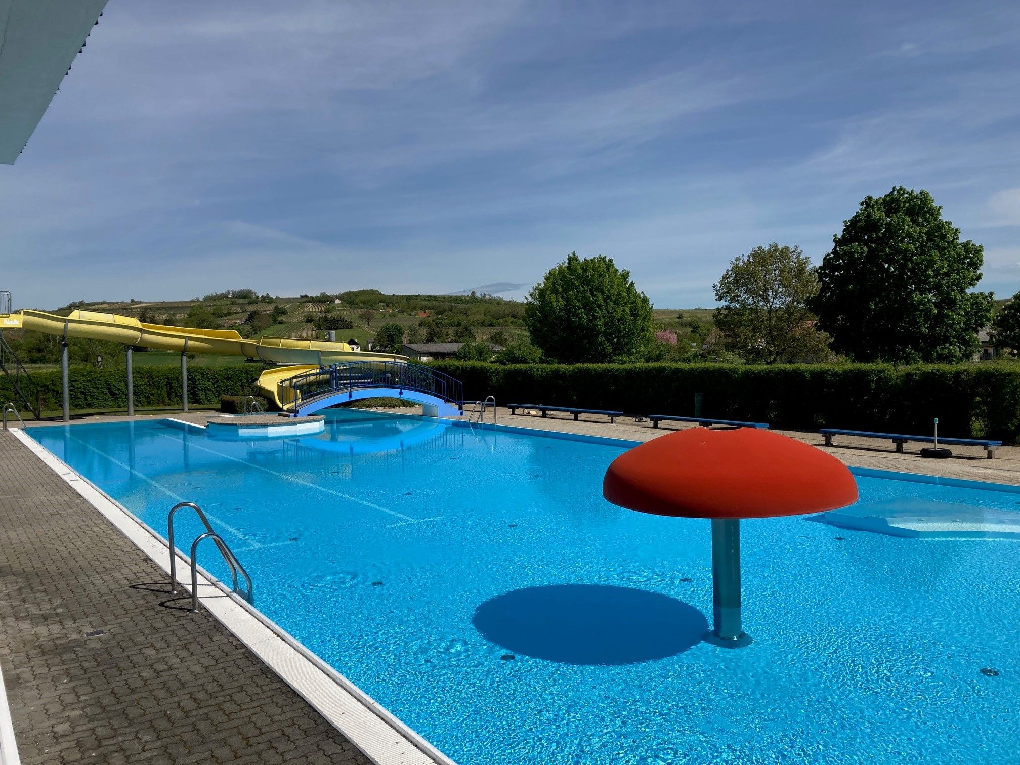 An outdoor pool with a yellow water slide, a blue pool and a red water feature in the shape of a mushroom.