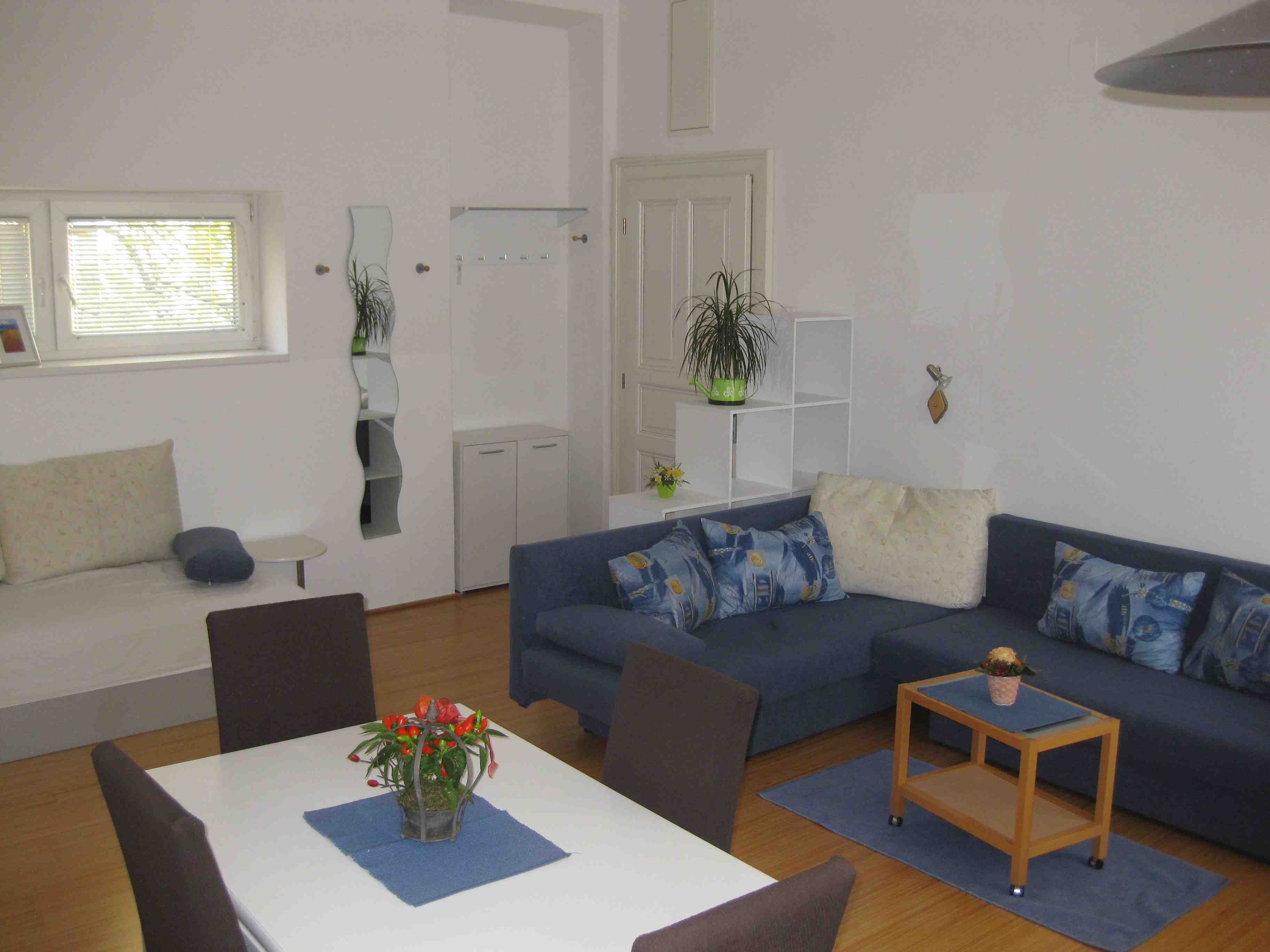 Apartment with blue sofa, white table and blue chairs. In the background a small window, door and mirror.