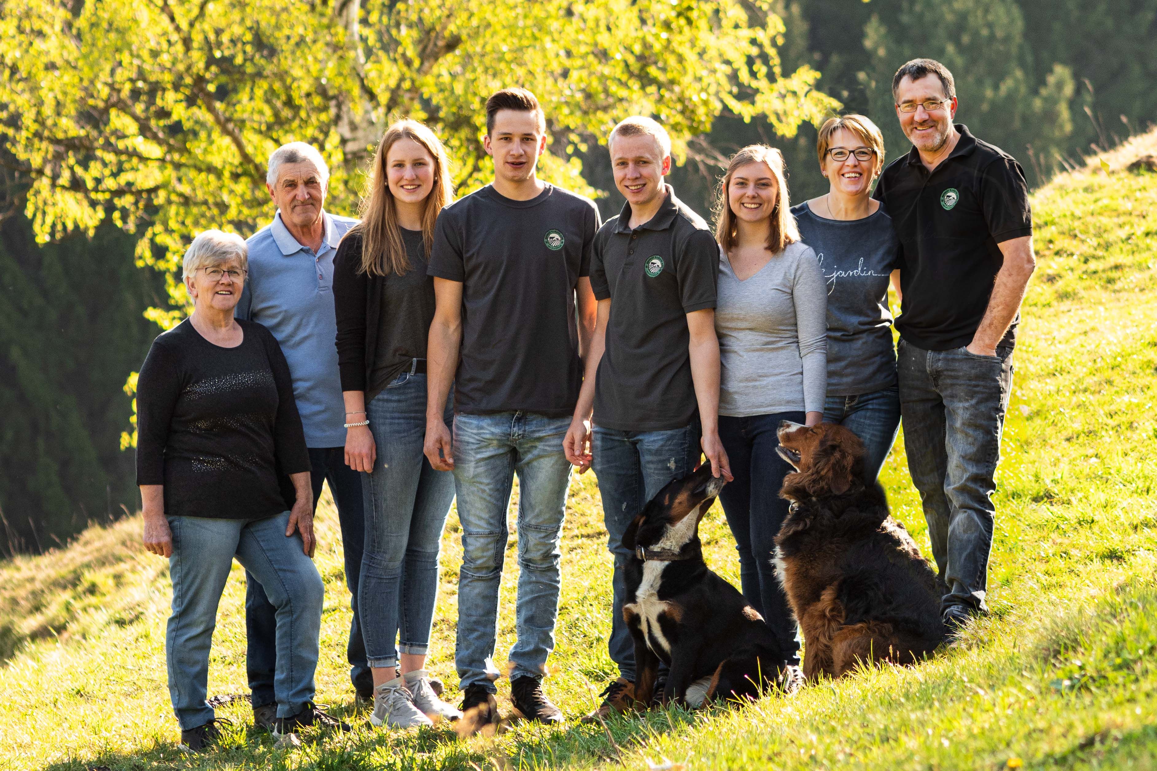 A large family with two dogs is standing on a green meadow in front of a tree.