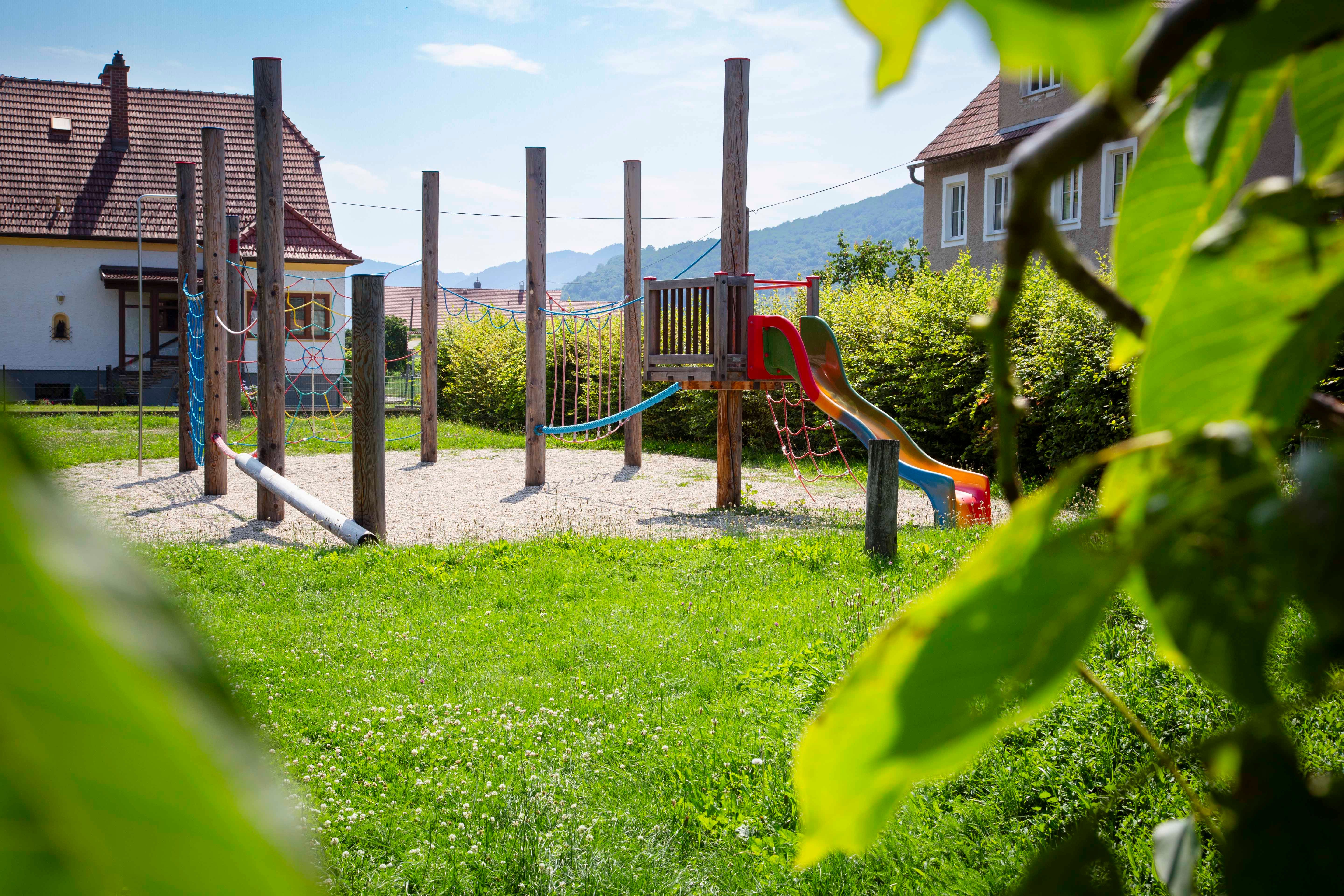 An adventure playground with climbing frame and slide, surrounded by green meadows and trees, with houses and hills in the background.